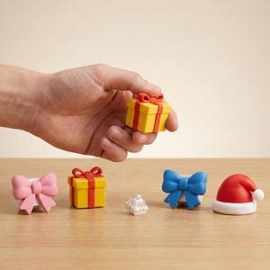 May include: A hand holding a small yellow gift box with a red bow. Other miniature items include a pink bow, another yellow gift box, a blue bow, and a Santa hat. All items are on a wooden surface.