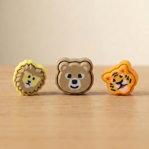 May include: Three animal-shaped erasers: a yellow lion, a brown bear, and an orange tiger. Each eraser features a cartoon-style face with black details. The lion has a yellow mane, the bear has a simple face, and the tiger has black stripes.
