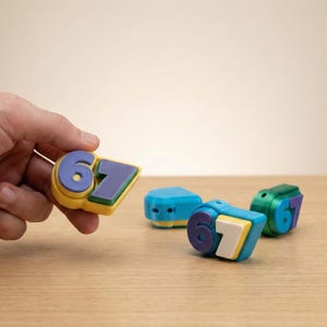 May include: A selection of brightly coloured number blocks, displaying the numbers 6 and 7. The blocks are in shades of blue, yellow, green, and white. One block is held in a hand, with others on a wooden surface.