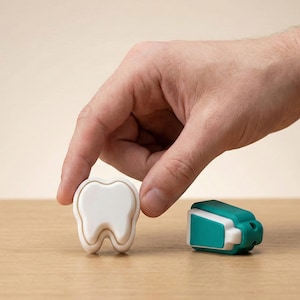May include: A white tooth-shaped object with a cream-coloured border and a teal and white dental floss dispenser on a wooden surface. A hand is reaching for the tooth-shaped object.