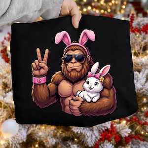 May include: Black tote bag featuring a cartoon Bigfoot wearing sunglasses and bunny ears, holding a white bunny. Bigfoot is making a peace sign with a pink peace sign bracelet. The bunny has a pink bow and flower.