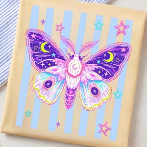 May include: A beige t-shirt featuring a graphic of a moth with purple, pink, and blue wings decorated with yellow crescent moons and stars. The moth is centered over a background of vertical light blue stripes and scattered pink and blue stars.
