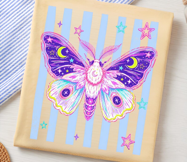 May include: A beige t-shirt featuring a graphic of a moth with purple, pink, and blue wings decorated with yellow crescent moons and stars. The moth is centered over a background of vertical light blue stripes and scattered pink and blue stars.