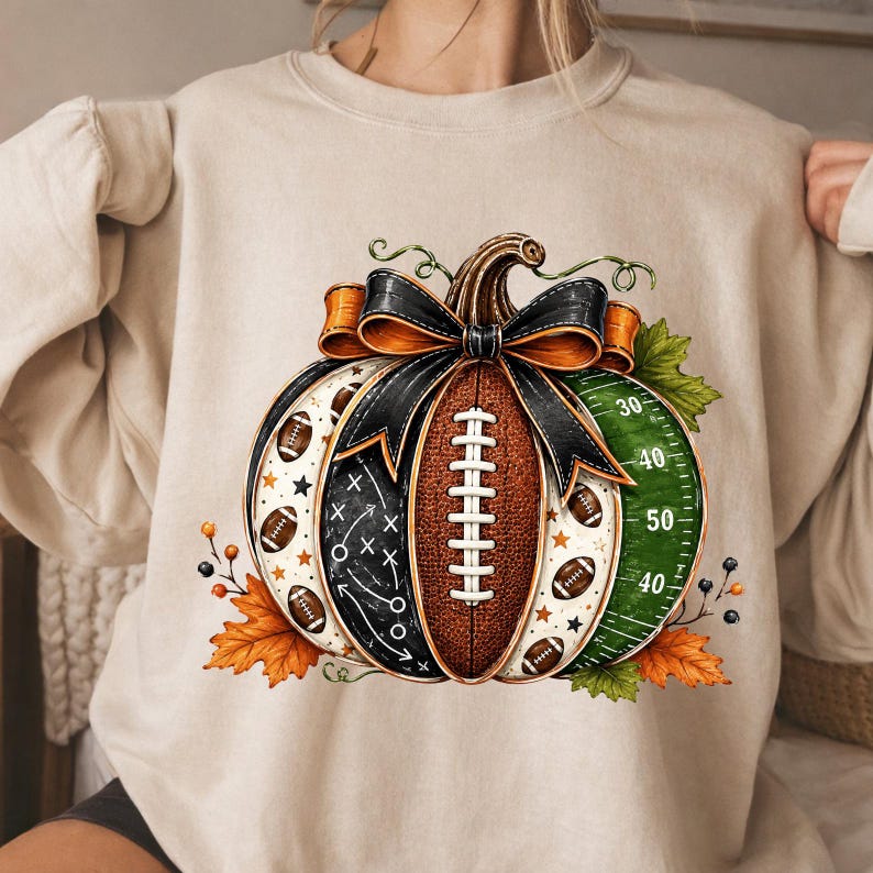 May include: A beige crewneck sweatshirt featuring a central graphic of a pumpkin designed with football-themed elements, including a brown football texture, a black chalkboard play diagram, and a green field pattern with white yard lines.