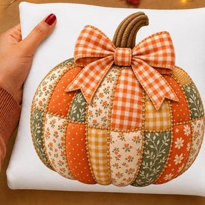 May include: A square white throw pillow featuring a central pumpkin graphic composed of various quilted fabric patches in orange, green, and cream tones. The pumpkin is adorned with a large orange and white gingham bow and a textured brown stem.