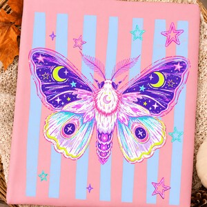 May include: A rectangular pink fabric item featuring vertical light blue stripes and a central graphic of a moth. The moth has purple, blue, and white wings decorated with yellow crescent moons and small stars, surrounded by scattered pink and blue stars.