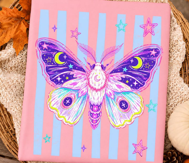 May include: A rectangular pink fabric item featuring vertical light blue stripes and a central graphic of a moth. The moth has purple, blue, and white wings decorated with yellow crescent moons and small stars, surrounded by scattered pink and blue stars.