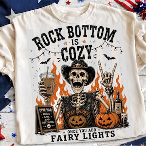 Peut inclure: Un t-shirt couleur crème avec un motif représentant un squelette portant un chapeau de cowboy et un t-shirt marqué STAY SPOOKY. Le squelette tient une boisson et fait un signe de la main rock. Le texte indique ROCK BOTTOM IS COZY ONCE YOU ADD FAIRY LIGHTS.