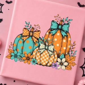 May include: A square pink canvas featuring a graphic illustration of three pumpkins. One teal pumpkin with orange polka dots, one orange pumpkin with white polka dots, and one checkered pumpkin. Each pumpkin is adorned with a decorative bow and flowers.
