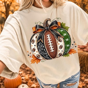 May include: A cream-colored t-shirt featuring a graphic of a pumpkin designed with football elements. The pumpkin includes a brown football texture, a black bow, a green field pattern with yard lines, and white play diagrams, accented by orange leaves.