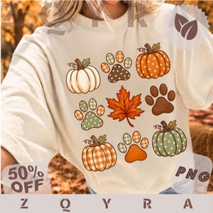 Puede incluir: Una camiseta de color crema con un estampado gráfico de tres calabazas, tres huellas de pata y una hoja de arce. Las calabazas y las huellas están decoradas con varios patrones, incluyendo cuadros vichy naranjas y blancos, lunares y colores sólidos.