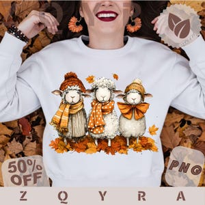 Op de afbeelding: Een wit sweatshirt met lange mouwen en een grafische illustratie van drie schapen die in herfstbladeren staan. De schapen dragen oranje en bruine gebreide mutsen en sjaals. Het middelste schaap draagt een sjaal met stippen.