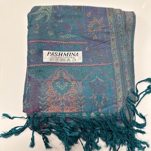 May include: A folded pashmina scarf with a teal, purple, and orange floral pattern. The scarf has a fringe along the bottom edge and a label that reads "PASHMINA 100% PASHMINA".