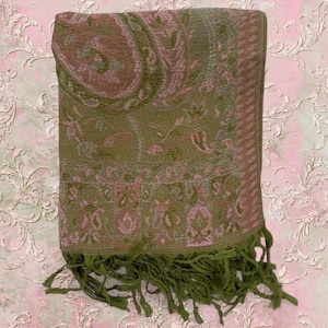 Può includere: Una sciarpa tessuta rettangolare con una base verde oliva e un motivo paisley ripetuto in tonalità rosa tenue e marrone spento. Il tessuto ha un aspetto strutturato ed è rifinito con lunghe frange verde oliva abbinate lungo il bordo inferiore.