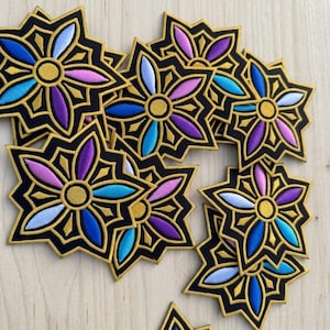 May include: A collection of star-shaped embroidered patches featuring a central gold circle surrounded by six petals in shades of blue, purple, and light pink. Each patch has a thick black border and a gold outline, arranged in a scattered pattern.