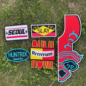 May include: A selection of embroidered patches with different designs and text. The patches feature the words "SEOUL", "SLAY", "HUNTRIX World Tour", and "Rrrrrrumi". The patches are in various shapes and colours, including red, black, yellow, and blue.