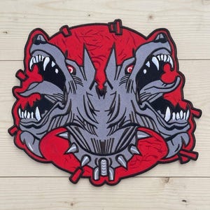 May include: An embroidered patch featuring a three-headed grey wolf creature with sharp white teeth and red eyes. The creature is set against a circular red background with black outlines and small red spikes extending from the edges of the design.