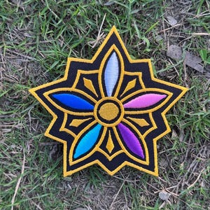 May include: Embroidered patch featuring a stylized flower design. The patch has a black background with a gold border and accents. The flower petals are in shades of blue, purple, pink, and white. The patch is set against a background of green grass.