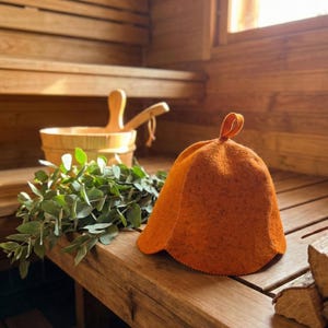 Peut inclure: Un chapeau de sauna en feutre orange est posé sur un banc en bois à l'intérieur d'un sauna. Un seau en bois avec une louche et un bouquet de branches vertes feuillues sont également sur le banc. L'intérieur du sauna est en bois clair.