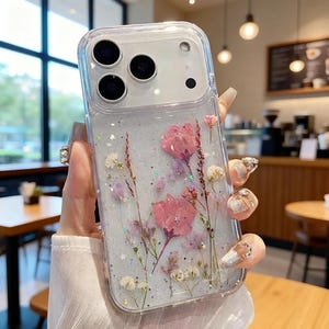 以下が含まれることがあります： ピンクと白のドライフラワーとグリッターが埋め込まれた透明な携帯電話ケース。ケースは透明なデザインで、携帯電話のカメラとフラッシュを際立たせています。花の配置には繊細な茎と花びらが含まれています。