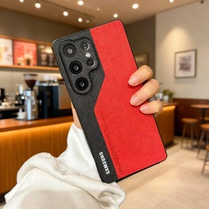 Puede incluir: Una funda roja y negra para teléfono Samsung. La funda tiene una sección negra texturizada en el lado izquierdo con las lentes de la cámara y una sección roja en el lado derecho. El logotipo de Samsung es visible en la parte inferior. La funda se sostiene en una mano.