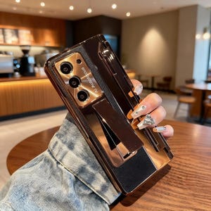 Puede incluir: Una funda de teléfono de oro rosa con un soporte incorporado. La funda tiene un acabado metálico y un acento negro similar al cuero. El área de la cámara es visible, con el texto "AI Autofocus" impreso en la funda.