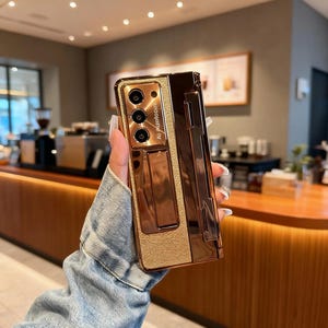 May include: A metallic copper-colored phone case with a textured panel and a built-in stand. The case has a glossy finish and features the text "AI Autofocus". The case is designed to protect a folding smartphone.
