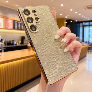 May include: Gold-colored phone case with a textured, wave-like pattern. The case has a gold frame and a camera module with multiple lenses. The phone case is designed to fit a specific phone model.