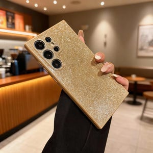 May include: A gold glitter phone case with a camera cutout. The case is rectangular and has a shiny, textured surface. The phone case is held by a person.