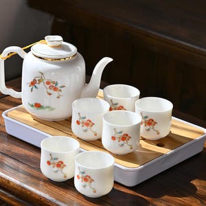 Peut inclure: Service à thé en porcelaine blanche comprenant une théière et six tasses, chacune ornée d'un motif floral rouge, orange et vert. La théière a un bord doré et repose sur un plateau en bambou. Pour le service du thé.