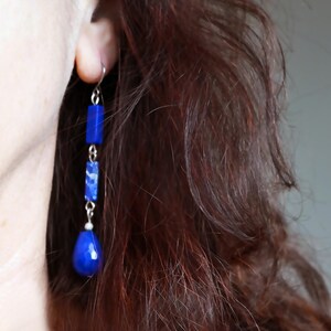 May include: A close-up of a person's ear wearing long, dangling earrings. The earrings feature a series of blue, rectangular and teardrop-shaped beads. The beads are strung together with silver-colored metal. The person has long, dark brown hair.