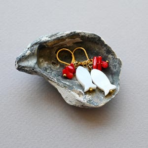 Può includere: Un paio di orecchini con pendenti bianchi a forma di pesce e perline di corallo rosso, esposti in un guscio di ostrica grigio. Gli orecchini hanno ganci color oro. Lo sfondo è grigio chiaro.