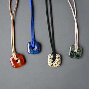 May include: Four square pendant necklaces with leather cords. The pendants are red, blue, black and green with unique patterns. The necklaces are displayed against a grey background.