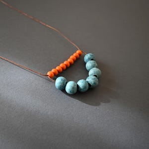 May include: A handmade necklace featuring a string of small orange beads and larger, round, blue-green beads. The beads are strung on a thin, brown cord, creating a unique and artistic design. The necklace is displayed against a gray background.
