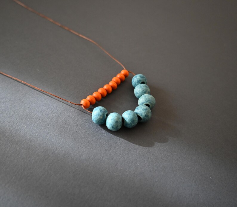 May include: A handmade necklace featuring a string of small orange beads and larger, round, blue-green beads. The beads are strung on a thin, brown cord, creating a unique and artistic design. The necklace is displayed against a gray background.