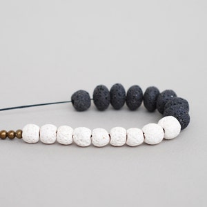 Op de afbeelding: Een armband van zwarte en witte lavasteen kralen met gouden kralen. De armband is gemaakt van natuurlijke lavasteen kralen en is geregen op een zwart koord.