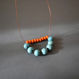 May include: A handmade necklace featuring a string of teal blue beads in a semi-circle, connected to a row of orange beads. The necklace is strung on a thin, brown cord. The beads have a matte finish and are set against a gray background.