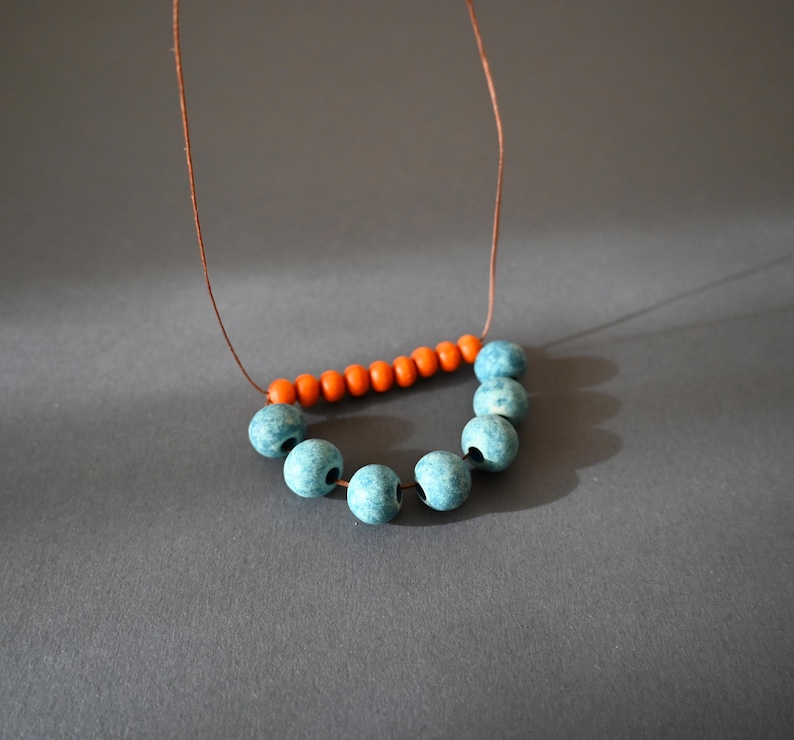 May include: A handmade necklace featuring a string of teal blue beads in a semi-circle, connected to a row of orange beads. The necklace is strung on a thin, brown cord. The beads have a matte finish and are set against a gray background.
