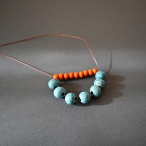 May include: A handmade necklace featuring a string of turquoise beads and a row of orange beads. The beads are strung on a thin, brown cord. The necklace is displayed on a gray surface.