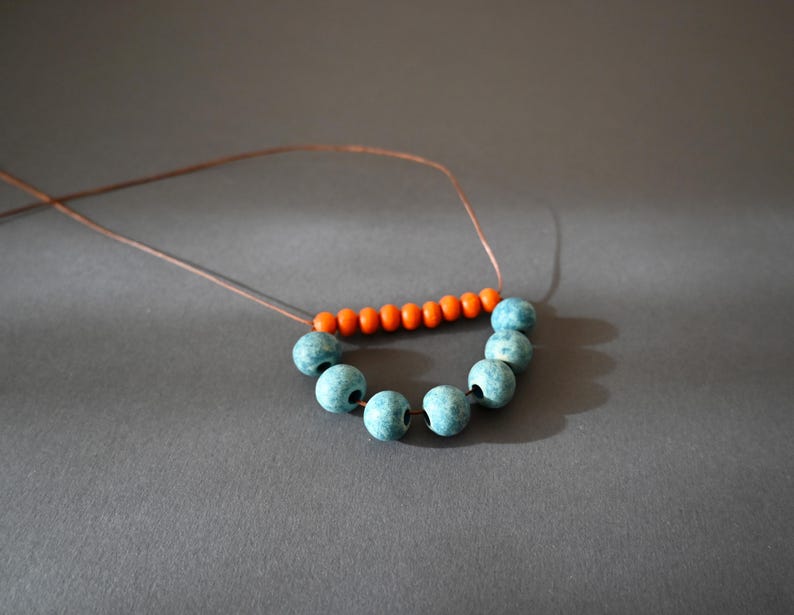 May include: A handmade necklace featuring a string of turquoise beads and a row of orange beads. The beads are strung on a thin, brown cord. The necklace is displayed on a gray surface.
