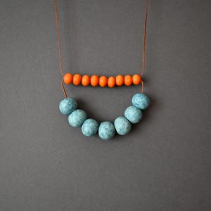 May include: A handmade necklace featuring a string of small orange beads above a curved row of larger, speckled blue beads. The necklace is displayed against a gray background, highlighting the contrasting colors and textures of the jewelry.