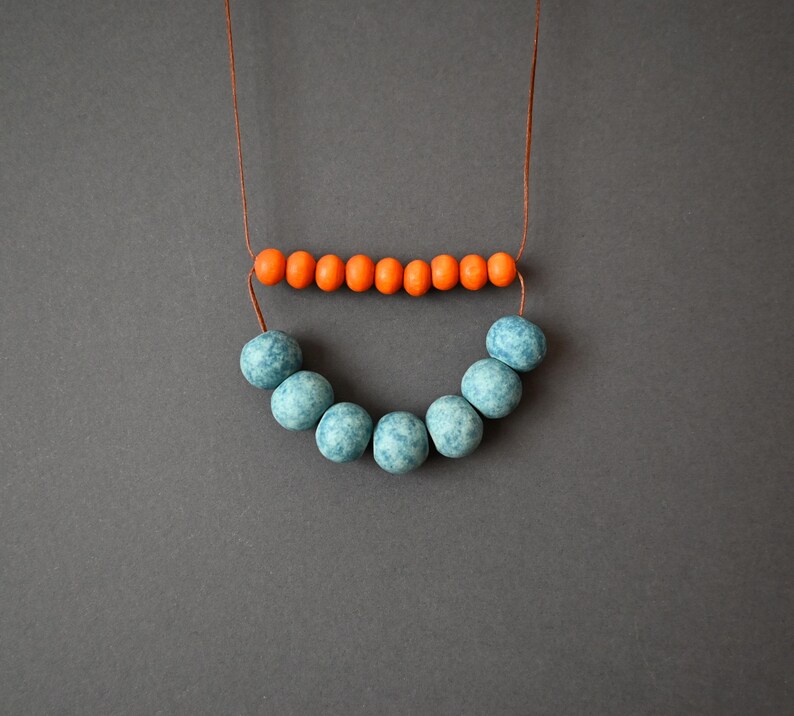 May include: A handmade necklace featuring a string of small orange beads above a curved row of larger, speckled blue beads. The necklace is displayed against a gray background, highlighting the contrasting colors and textures of the jewelry.