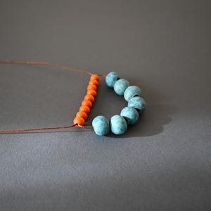 May include: A necklace in progress, featuring orange and turquoise beads strung on a brown cord. The orange beads are arranged in a straight line, while the turquoise beads form a curved shape. The background is a solid gray.