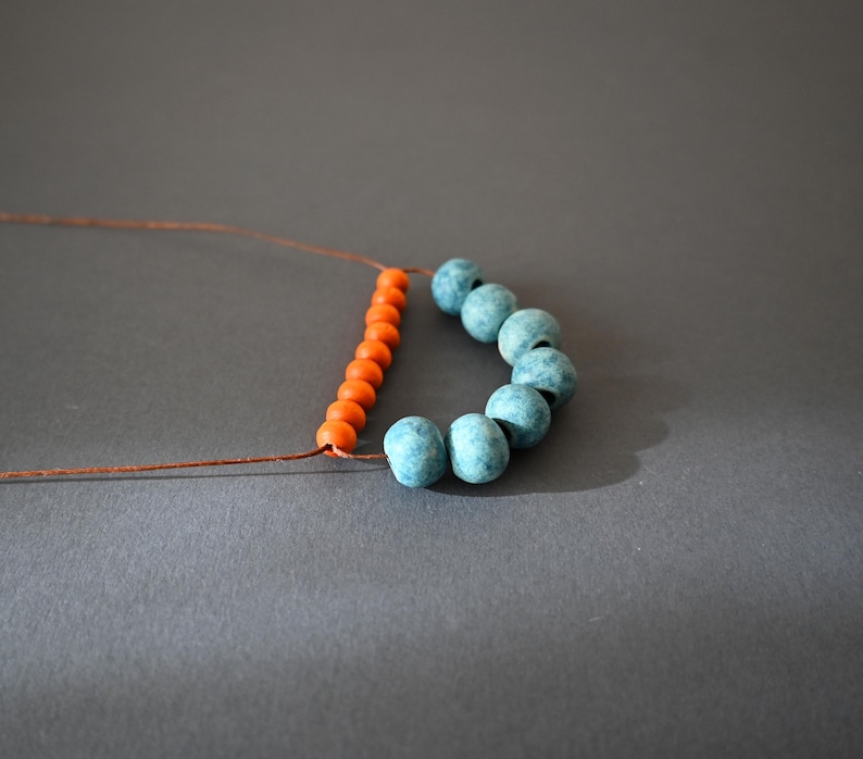 May include: A necklace in progress, featuring orange and turquoise beads strung on a brown cord. The orange beads are arranged in a straight line, while the turquoise beads form a curved shape. The background is a solid gray.