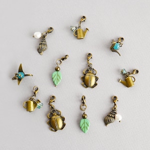 Op de afbeelding: Een verzameling bronzen bedels met verschillende ontwerpen, waaronder een schelp met een parel, een gieter, een kever, een blad en een vogel. Sommige bedels hebben turquoise of heldere kristallen accenten. De bedels zijn ontworpen om aan armbanden of kettingen te worden bevestigd.