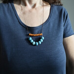 May include: A necklace with a brown cord featuring two rows of beads. The top row has small orange beads, and the bottom row has larger, light blue beads. The necklace is worn against a dark blue t-shirt.