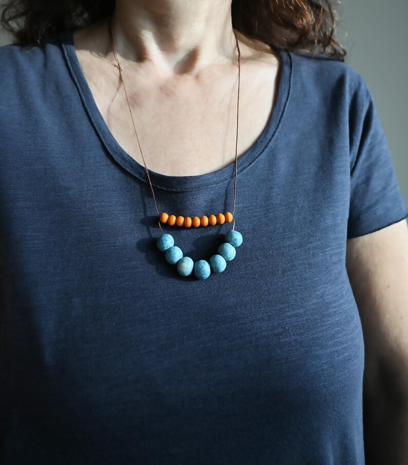 May include: A necklace with a brown cord featuring two rows of beads. The top row has small orange beads, and the bottom row has larger, light blue beads. The necklace is worn against a dark blue t-shirt.