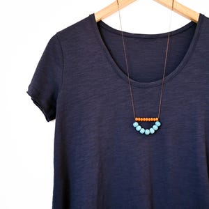 May include: A navy blue scoop-neck t-shirt hangs on a wooden hanger. A necklace with a brown cord features a row of orange beads above a curved row of light blue beads, creating a minimalist design.