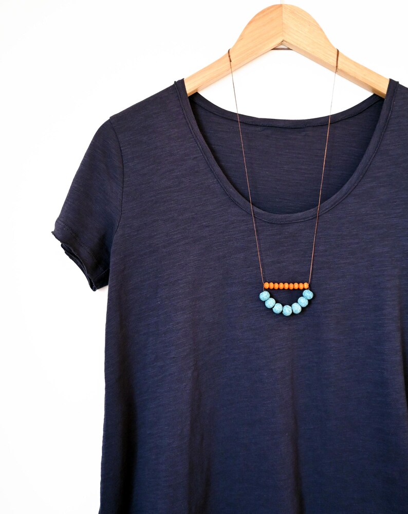 May include: A navy blue scoop-neck t-shirt hangs on a wooden hanger. A necklace with a brown cord features a row of orange beads above a curved row of light blue beads, creating a minimalist design.
