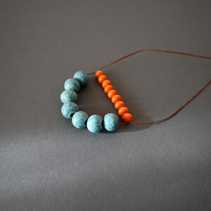 May include: A necklace featuring a combination of blue and orange beads. The blue beads are clustered in a curved line, while the orange beads form a straight line. The beads are strung on a thin, brown cord, creating a simple yet striking design.
